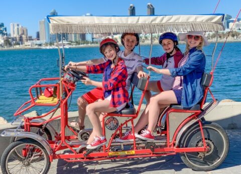 Family riding in a red six person surrey rented from Wheel Fun Rentals on Coronado Island. San Diego skyline in the backdrop