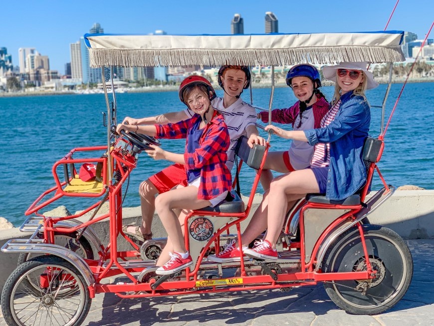 Family riding in a red six person surrey rented from Wheel Fun Rentals on Coronado Island. San Diego skyline in the backdrop