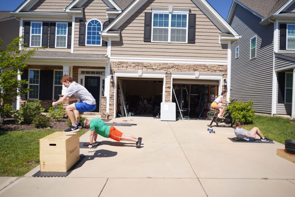 Family working out at various stations set up in driveway in front of house. Get the whole gang moving with a series of exercise stations like jump rope, burpees, and ab mat sit-ups. 