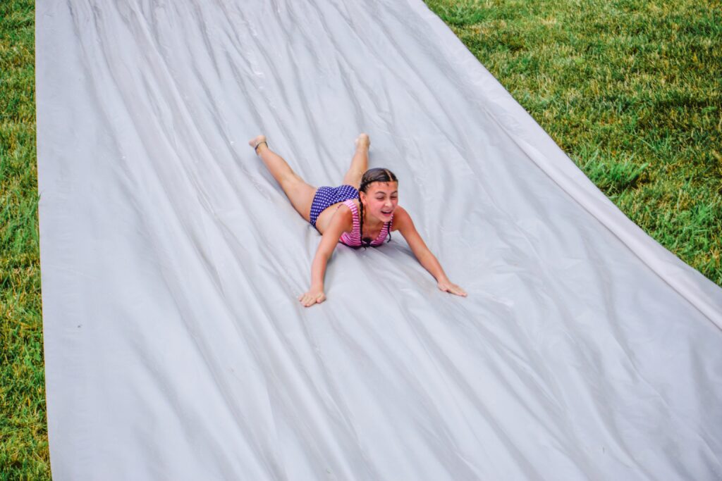 Girl slides down a large slip and slide. Slip ‘n slides are a fun way to cool off on a hot summer day, no matter what age you are. Try making your own with a large roll of clear or white heavy duty plastic sheeting so that it doesn’t get hot and landscaping stakes to hold it in place. Just add water and some Dawn dish soap and you are ready to go! - Safe Summer Bucket List – 100 Ways to Have Fun This Summer While Social Distancing