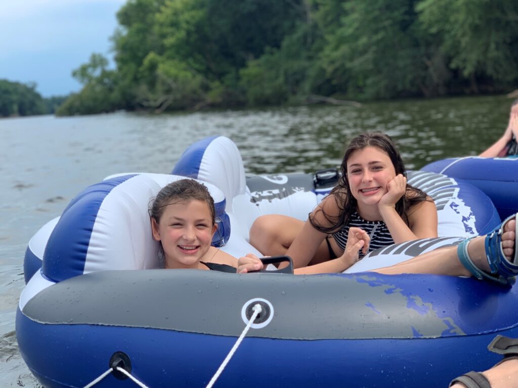 Two girls spend a relaxing summer day slow tubing down a river in rafts. - Safe Summer Bucket List – 100 Ways to Have Fun This Summer While Social Distancing