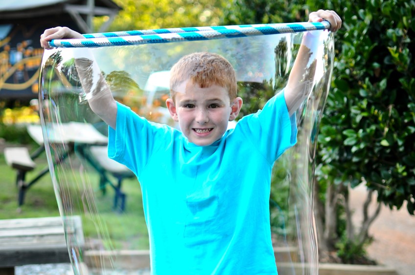 Young boy stands inside giant bubble made with a hula hoop. Try using a kiddie pool and hula hoop to make bubbles big-enough your kids can fit inside them. 