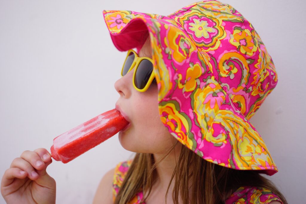 Girl in colorful hat and sunglasses enjoys strawberry popsicle. On a hot summer day, fresh-from-the-freezer homemade popsicles really hit the spot. 