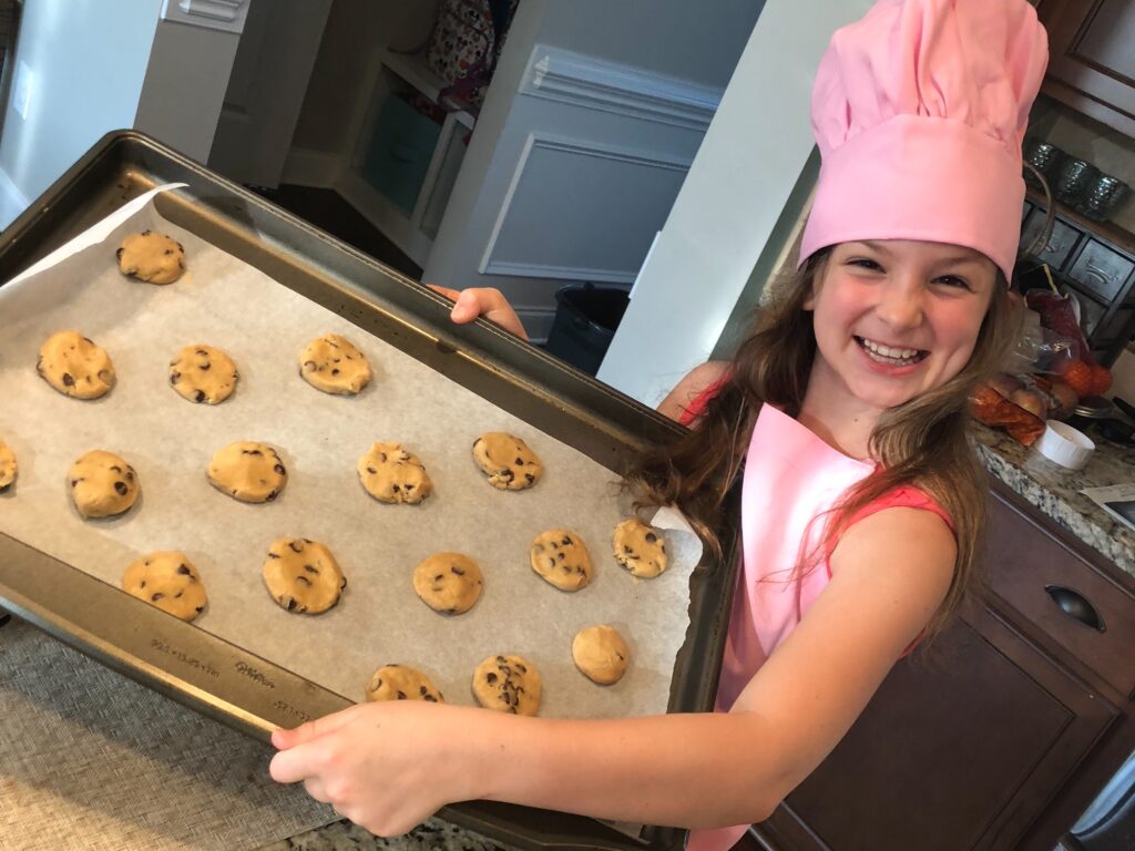 Smiling girl holding pan of cookie dough ready to go in the oven. Get out those cook books and try baking something you’ve never made before.- Safe Summer Bucket List – 100 Ways to Have Fun This Summer While Social Distancing