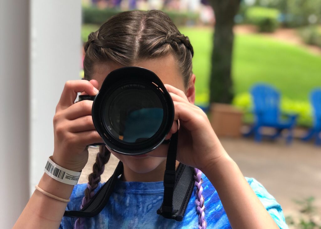 Young girl holds DSLR camera up to her face. No doubt you will want to capture all the awesome memories you will be making over the summer together. Now is the perfect time to get your kids behind the camera too and teach them the ropes of photography. Check out my tips for taking better family photos to get you started. -No doubt you will want to capture all the awesome memories you will be making over the summer together. Now is the perfect time to get your kids behind the camera too and teach them the ropes of photography. Check out my tips for taking better family photos to get you started. - Safe Summer Bucket List – 100 Ways to Have Fun This Summer While Social Distancing