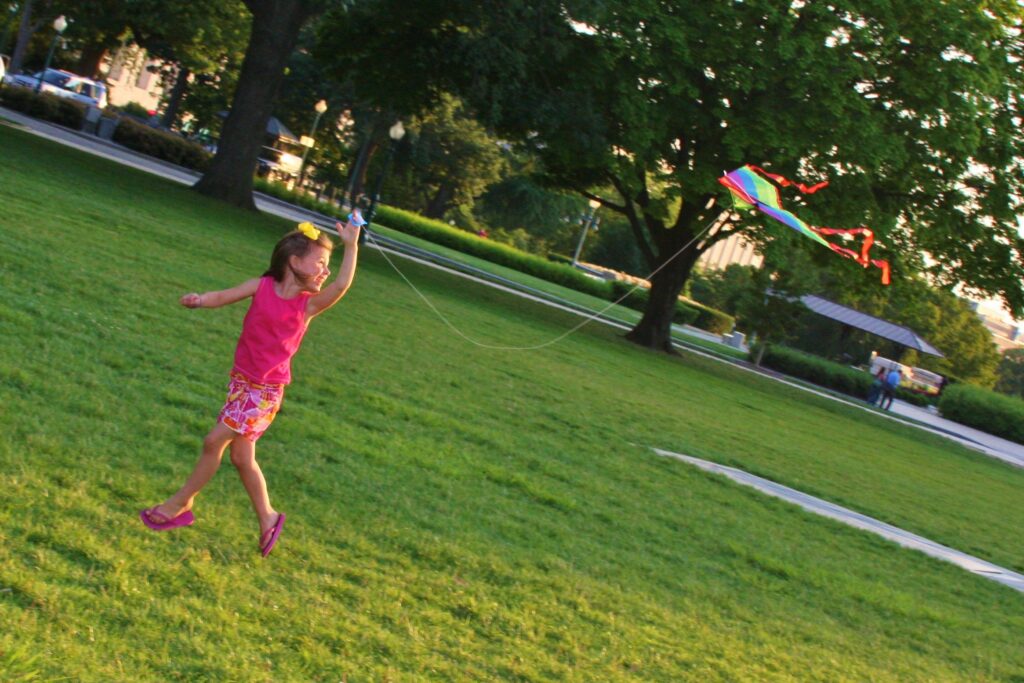 Young girl flies a rainbow kite in an open field. - Safe Summer Bucket List – 100 Ways to Have Fun This Summer While Social Distancing