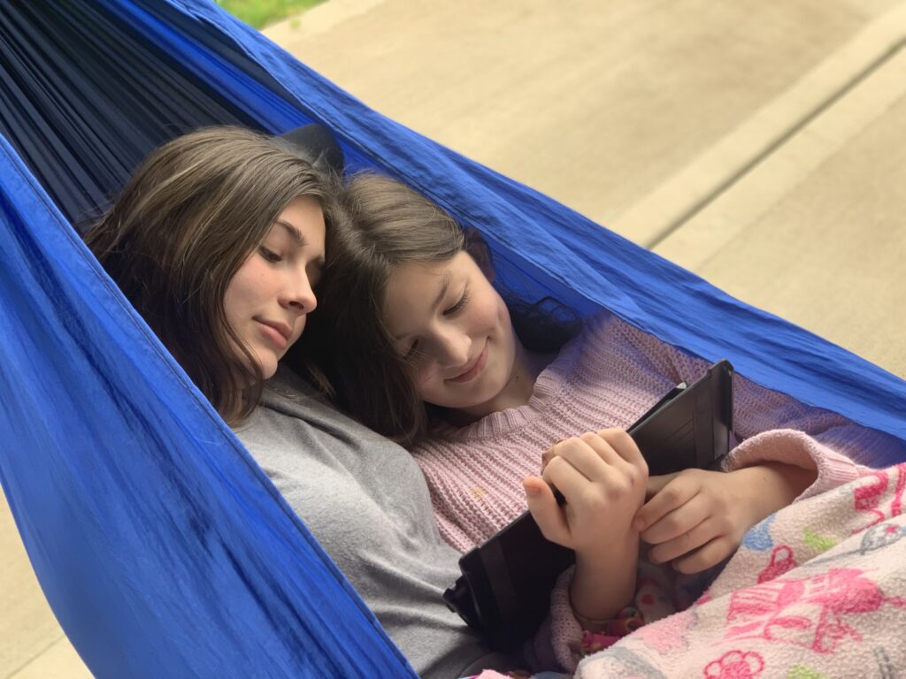 Two girls hanging out in a hammock watching ipad. - Safe Summer Bucket List – 100 Ways to Have Fun This Summer While Social Distancing