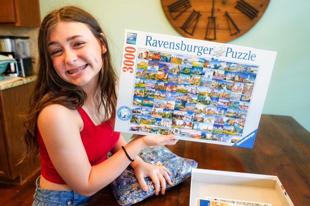 Teen Girl holds up giant 3000 piece jigsaw puzzle. Now is the perfect time to try a challenging puzzle that will take at least a couple of days to complete. Set it up on your dining room table or find an empty spot where you can all work together. - Safe Summer Bucket List – 100 Ways to Have Fun This Summer While Social Distancing