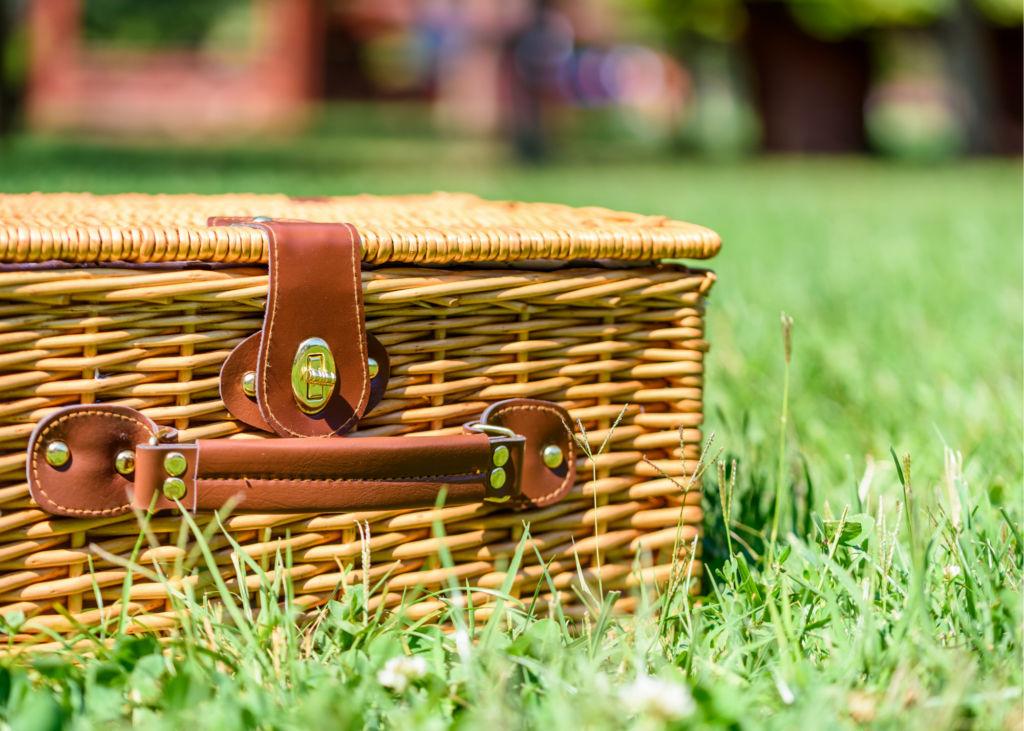 Image of a wicker picnic basket in the grass. Complete with a blanket and basket, enjoy a picnic in your backyard, or a secluded spot with a special view. Let your kids plan the menu and pack everything up. - Safe Summer Bucket List – 100 Ways to Have Fun This Summer While Social Distancing