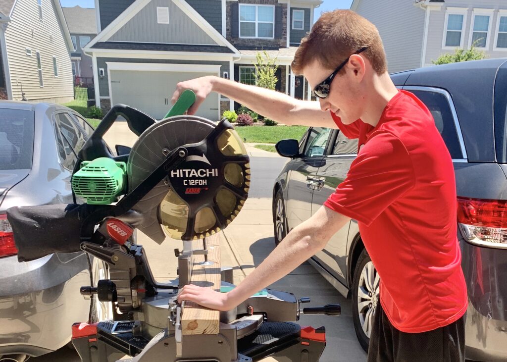 Teenage boy uses saw to cut wood. This summer is the perfect time to learn to safely use power tools. Create a corn hole set, build a raised bed garden or a new home for Fido. Look online for free build plans and what tools you will need. 