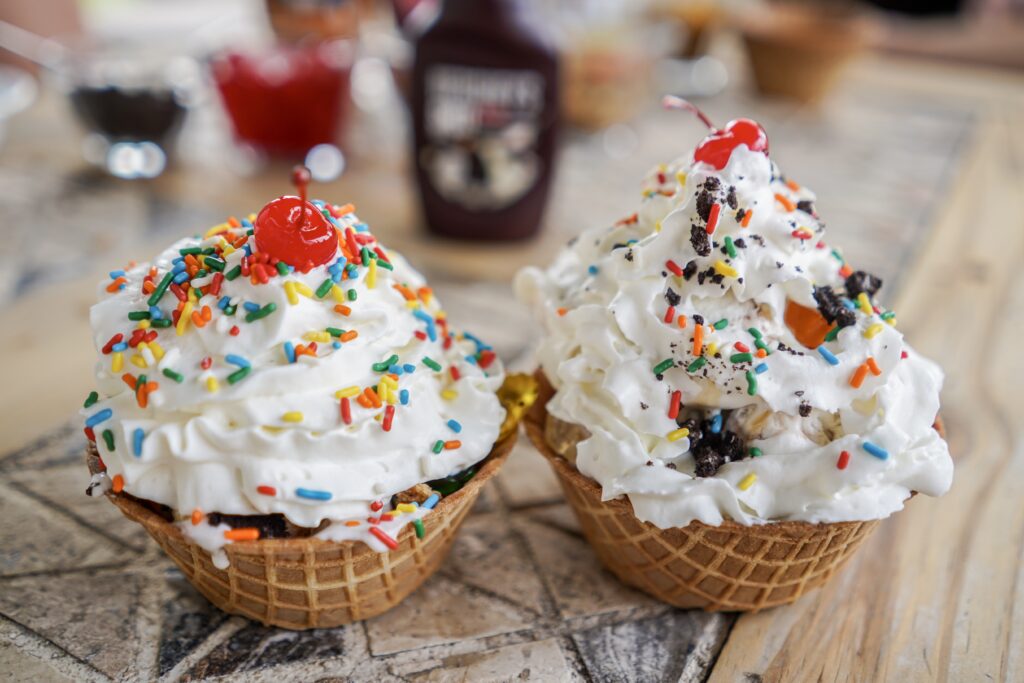 Two ice cream sundaes in waffle bowls, topped with whip cream, cherry and sprinkles. Create an ice cream bar with options for sundaes, ice cream sandwiches, toppings and more. You can even make your own home-made ice cream using a counter-top ice-cream machine or rock-salt , zip-lock bags and ice. - Safe Summer Bucket List – 100 Ways to Have Fun This Summer While Social Distancing